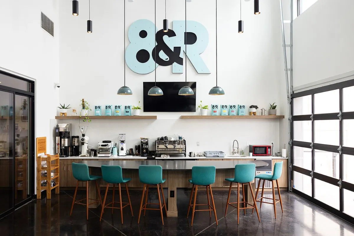 The inside of a coffee shop with white walls and bright teal stools at a counter